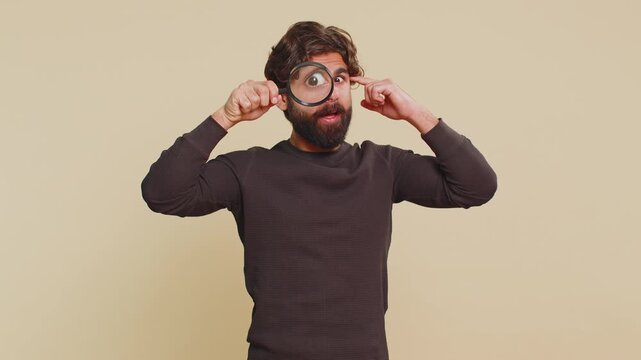 Investigator researcher scientist man holding magnifying glass near face, looking into camera with big zoomed funny eyes, searching, analyzing. Young Hindu guy isolated on beige studio background.