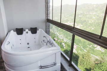 Obraz premium Modern white jacuzzi bathtub placed on a glass enclosed balcony with panoramic mountain valley view, symbolizing luxury wellness, spa relaxation, travel comfort and peaceful nature retreat lifestyle 