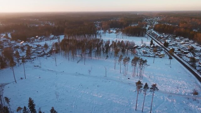 Aerial view shows snow fields, pine trunks, a straight road, and small houses. Warm low sun casts long orange highlights and shadows. Slow pan and drift reveal details.