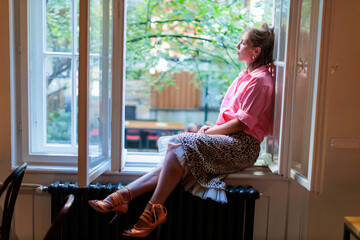 Woman sits on a windowsill with her legs crossed while looking outside at a bright sunny day