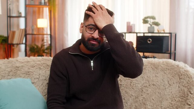 Indian man at home sits on sofa and covers face with hand in frustration after realizing mistake. Arabian guy reacts with facepalm gesture, showing emotional disappointment and regretful reaction.