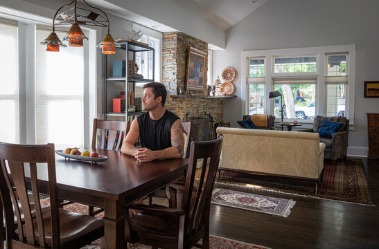 Young strong athletic man with tattoos at a dining room table inside a house. 