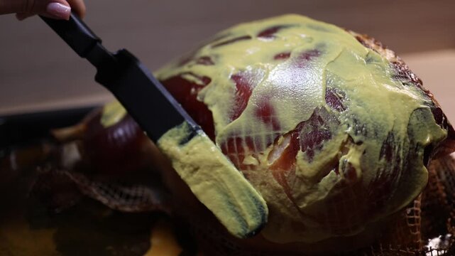 Roasted pork knuckle on baking tray as mustard is spread on top with spatula close up festive dinner preparation showing seasoning process rich texture and home cooking atmosphere