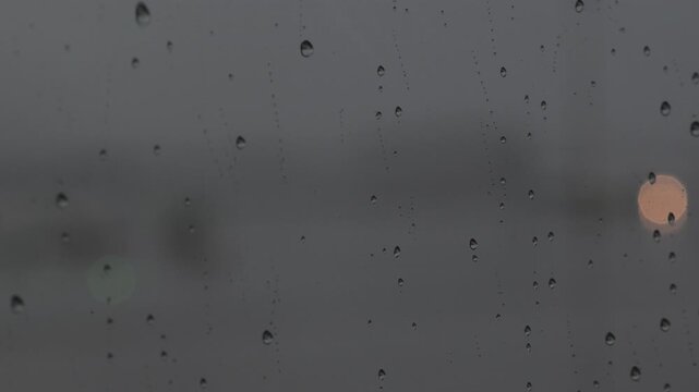 Rain falls on a window, creating drops and streaks during a cloudy day in a quiet city