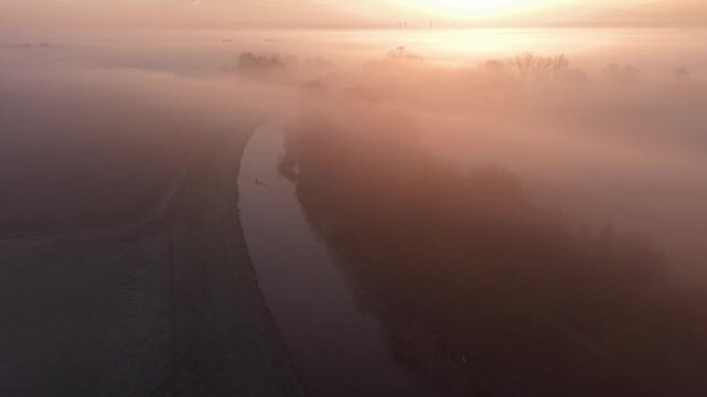 Aerial view of foggy canal at sunrise, flanked by trees and fields. Wind turbines silhouette the horizon as warm light diffuses through mist, evoking serenity, sustainability, and rural elegance.