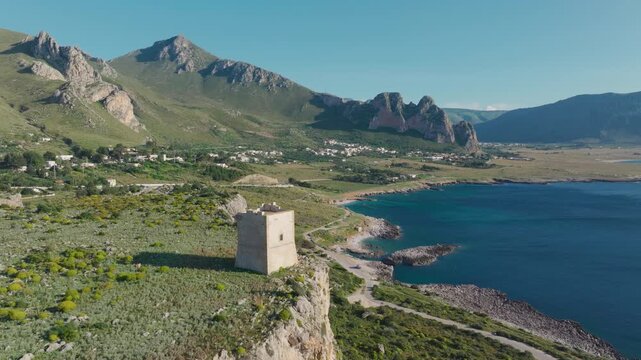 AERIAL: Historic stone watchtower perched on sea cliff above winding coastal road. Turquoise sea bordering a rugged coastline, mountains and a small village spread out near San Vito Lo Capo in Sicily.
