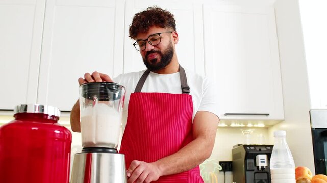 Arabian man blending whey protein smoothie with banana apple after workout in home kitchen. Young guy smiling using blender for healthy sports nutrition drink for muscle recovery in small apartment.