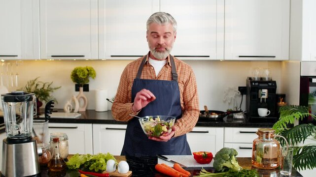 Middle-aged man presenting fresh vegetable salad in bowl smiling while filming pro cooking tutorial for clean eating. Mature guy sharing diet meal prep tips for nutrition and wellness in home kitchen.