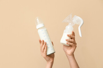Female hands holding breast pump and bottle of milk on beige background