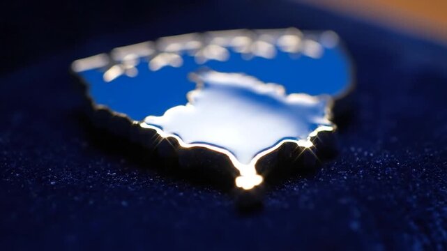 A macro shot of a glossy blue enamel map-shaped lapel pin resting on dark blue velvet. Subtle movements and lighting shifts highlight the silver trim and reflective surface of the keepsake.