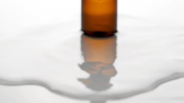 An amber dropper bottle settles into a pool of white liquid on a reflective surface, transitioning from initial splash and ripples to a calm, smooth puddle, highlighting cosmetic or wellness liquid f