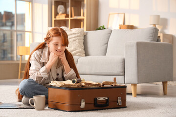 Cute teenage happy redhead girl with adventure book, world map and suitcase sitting on floor at home