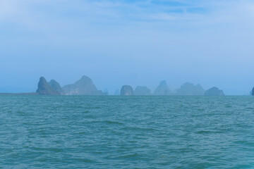 A tropical island in the blue sea features a rocky mountain coastline and calm ocean water under a cloudy summer sky in a beautiful Thailand landscape