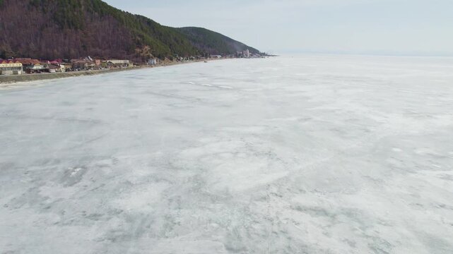Flight over the ice of Lake Baikal in spring, the village of Listvyanka. Cars with tourists are driving along the road along the lake, and hovercraft are driving on the ice.