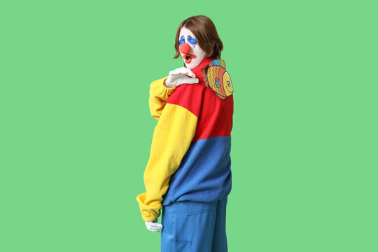 Young shocked woman dressed as sad clown with paper fish attached to her back on green background. April Fool's day celebration