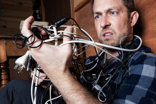 A stressed adult male struggles with a chaotic tangle of various electronic cables, USB cords, and chargers in a dark room. Conceptual image for technology frustration, IT problems, and the need for o