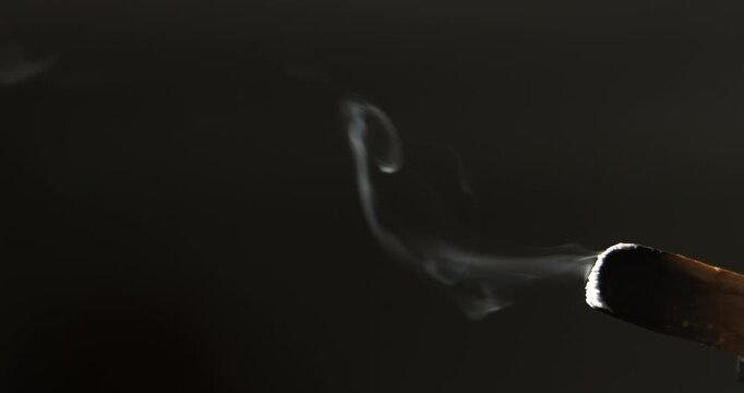 Swirling patterns of smoke drift from a glowing palo santo stick, highlighted by angled sunlight in a dark environment. 
