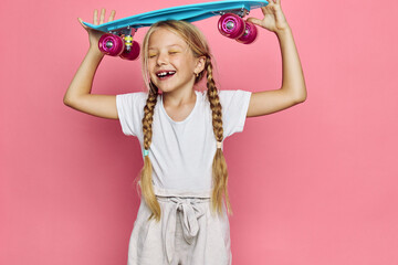 Happy young girl with braids holds a blue skateboard above her head with eyes closed. She smiles wearing a white t-shirt and light pants against a pink background. © SHOTPRIME STUDIO