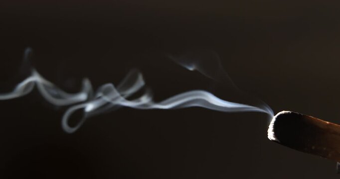 Swirling patterns of smoke drift from a glowing palo santo stick, highlighted by angled sunlight in a dark environment. 