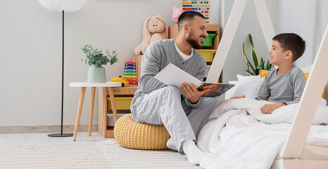 Naklejka premium Happy father with his son in pajamas reading book on bed at home