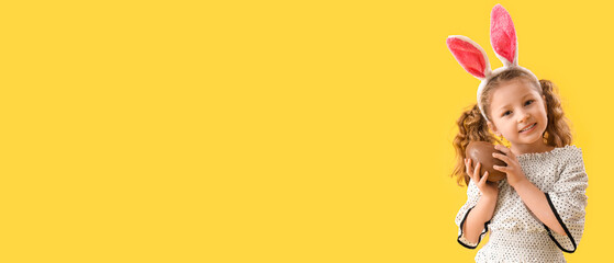 Cute little girl in bunny ears with chocolate Easter egg on yellow background