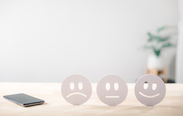 Fototapeta premium Online customer review satisfaction feedback on mobile phone app for user experience survey showing sad neutral happy face emojis on light wood table