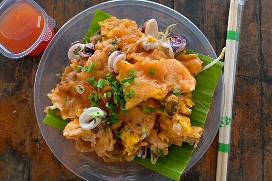 A savory dish of fried seafood omelette (Hoi Tod) with bean sprouts on a banana leaf, served with a tangy dipping sauce, at a food stall in Thailand