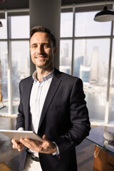 Positive young Hispanic executive man holding tablet, posing in modern office with large windows, looking at camera, smiling, using digital pad for work financial transaction. Vertical shot