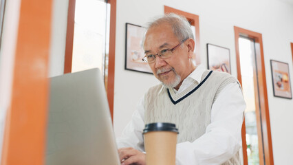 An elderly Asian man wearing glasses and a sweater vest focuses on his laptop while sitting in a...