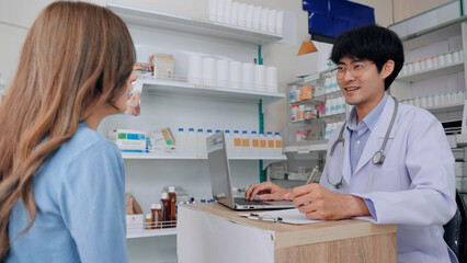 Fototapeta premium A professional Asian male pharmacist in a white lab coat consults a female patient in a modern drugstore. This medical depicts expert healthcare service and patient care