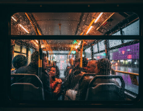Bus window is streaked with raindrops, obscuring the view of chatty passengers inside. Outside, the city's night comes alive with colorful lights, creating a warm and bustling atmosphere as rain gentl
