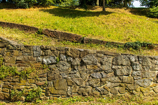 Old stone retaining wall in a public park with green grass and trees on a sunny day
