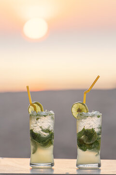 Two cold, steamed glasses of mojito cocktail with mint on a table in close-up on the beach, against the background of the setting sun at sunset, sand and sea. Vacation and travel concept lifestyle