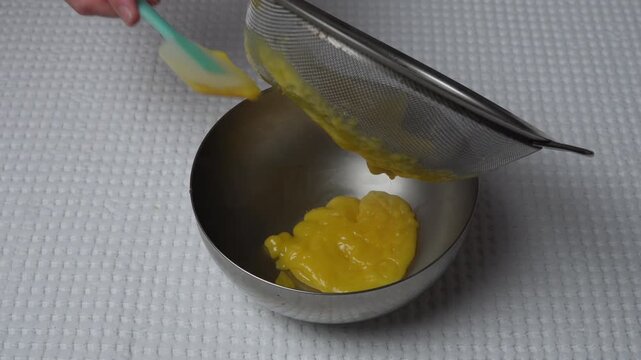 Scraping lemon curd from sieve into bowl. Hands scraping lemon curd from a sieve into a stainless steel bowl with a silicone spatula. Close-up finishing step of dessert preparation.