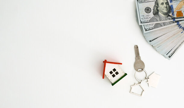 A model house, keys, and a stack of dollar bills on a white background with a place for text, top view. Concept for buying or renting real estate