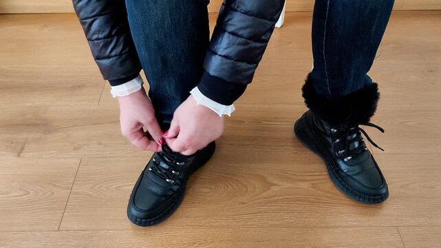 A figure sits on a wooden floor, tying the laces of black fur - lined winter boots - capturing a quiet, tactile moment of seasonal prep, warmth, and everyday cold - weather readiness
