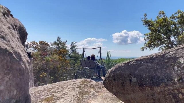 Anonymous couple sitting on a swing placed on rocky terrain in the La Cabrera mountains near Madrid, Spain, enjoying nature, open views, and a peaceful outdoor moment surrounded by trees and blue sky