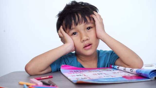 Bored Asian boy sitting at a desk with school books and crayons, looking tired and frustrated while doing homework. Stressful childhood education and learning difficulties concept in classroom.