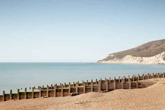 Coastal Beach Scene With Wooden Groyne, Pebble Shoreline, And White Cliffs Under Clear Sky