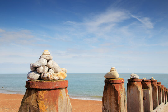 Stacked Stone Cairns On Rusty Seawall Posts Along A Calm Beach With Blue Ocean Sky