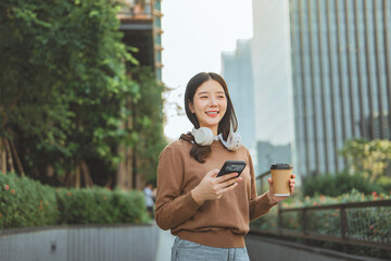 Happy Asian woman walking in a city business district, holding a coffee cup and smartphone with headphones around her neck. This concept represents modern urban lifestyle, travel, City life