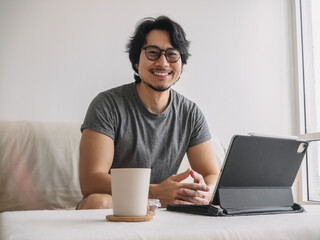 Happy Asian man smiling while working online with digital tablet computer in the living room, enjoy his work from home.