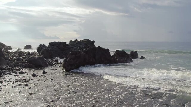 Aerial orbit shot of massive dark volcanic boulders on a pebbly shore with white ocean waves crashing under a cloudy sky in Flores, Indonesia.