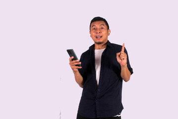 A happy looking Asian man wearing a blue shirt with a white undershirt points at an empty space while holding a cell phone, isolated on a white background.