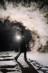 Obraz premium Individual in black coat creates smoke effect in snowy landscape at night, illuminated by a bright light source, with swirling vapor and snowflakes visible