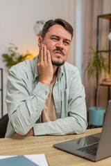 Naklejka premium Caucasian young man freelancer sitting at home office table feeling toothache touching mouth with hand, painful expression because of dental illness. gum disease, dental disease concepts. Vertical.
