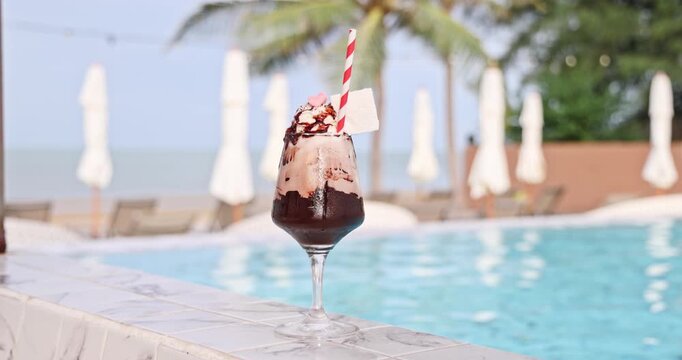 Chocolate milkshake with whipped cream, chocolate drizzle, chilling by a luxurious resort swimming pool, with palm trees and beach umbrellas in the background. Summer refreshment drink
