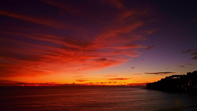 A breathtaking sunset paints the sky in vivid hues of orange and blue over a coastal town, with lush green hills framing the scene. A distant hilltop structure adds a serene focal point