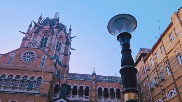 A lone street lamppost stands guard as the sunset bathes the Chhatrapati Shivaji Maharaj Terminus in a warm, ethereal glow, highlighting intricate Victorian architecture against a purple twilight sky.