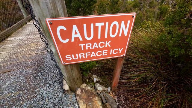 A slow camera push-in reveals a &ldquo;Caution Ice Surface&rdquo; warning sign, with a footbridge and surrounding wilderness visible in frame. The scene communicates environmental hazard, winter safety, outdoors.